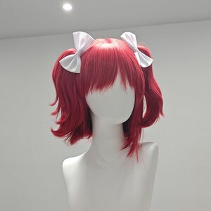 Puede incluir: Peluca roja con flequillo y dos lazos de satén blanco. La peluca está peinada con dos coletas bajas. La peluca se exhibe en una cabeza de maniquí blanca. El cabello es de un tono rojo vibrante.