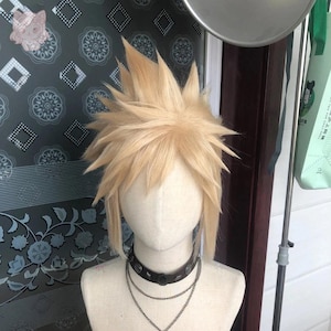 Könnte beinhalten: Blonde Cosplay-Perücke mit stacheligem, strukturiertem Haar, auf einem Schaufensterkopf gestylt. Die Perücke ist mit einem schwarzen Choker und einer mehrschichtigen Halskette kombiniert. Der Hintergrund zeigt eine gemusterte Wand.