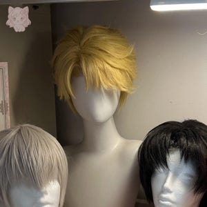 Peut inclure: Présentation de trois perruques sur des têtes de mannequin. La perruque centrale est d'un jaune doré clair avec un style en couches et une frange. Les deux autres perruques sont gris clair et noires. Les mannequins sont sur une étagère.