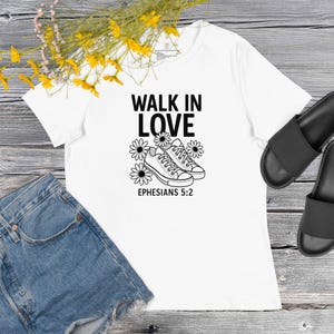 Puede incluir: Camiseta blanca con el texto "WALK IN LOVE" sobre un gráfico de zapatillas y margaritas. El texto "EPHESIANS 5:2" está debajo. La camiseta se combina con pantalones cortos de mezclilla y sandalias negras.