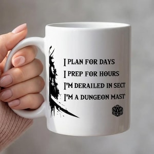 May include: White ceramic mug with black text that reads "I PLAN FOR DAYS, I PREP FOR HOURS, I'M DERAILED IN SECT, I'M A DUNGEON MAST." A black graphic of a sword and a dice are also on the mug. The mug is held by a person.