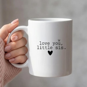 May include: White ceramic mug with a black heart and the text "love you, little sis." The mug has a curved handle and is held by a person wearing a pink sweater. The mug is a standard size.