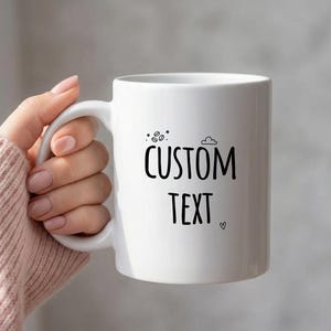 Op de afbeelding: Witte keramische mok met een gebogen handvat. De mok heeft de woorden "CUSTOM TEXT" in een zwarte, handgeschreven lettertype, met decoratieve elementen. Onder de tekst is een hart getekend.