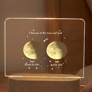 May include: A personalized night light with a wooden base and a clear acrylic panel. The panel features two moon illustrations, names, and dates, with the text "I love you to the moon and back." It has a simple on/off switch.