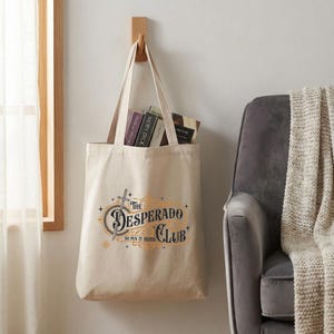 Puede incluir: Una bolsa de mano color crema con el texto "The Desperado Club" en un diseño decorativo, colgada de un gancho de madera. La bolsa contiene varios libros. Un sillón gris y un marco de ventana están en el fondo.