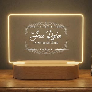 May include: A personalized LED desk lamp with a rectangular, illuminated acrylic panel. The panel displays the name "Jace Ryden" and "EVENT COORDINATOR" in elegant script within a decorative frame. The base is made of light-colored wood.