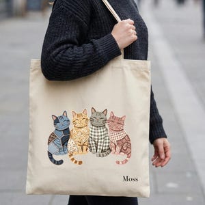 Könnte beinhalten: Beigefarbene Tragetasche mit vier stilisierten Katzen mit einzigartigen Mustern und Farben. Die Katzen sind nebeneinander angeordnet, jede mit einem unverwechselbaren Design, darunter Denim, Blumenmuster, Karomuster und Paisley. Das Wort "Moss" ist unten aufgedruckt.