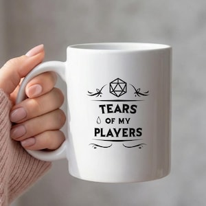 Op de afbeelding: Witte keramische mok met een zwarte afbeelding met de tekst "TEARS OF MY PLAYERS". De mok heeft een geometrische vorm en decoratieve krullen. De mok wordt door een persoon vastgehouden.