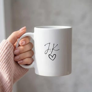 Peut inclure: Mug en céramique blanche avec une anse incurvée. Le mug présente les initiales « J.K » dans un style manuscrit, suivies d'un petit symbole de cœur. Le mug est tenu par une personne portant un pull rose.