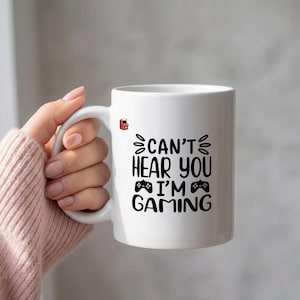 May include: White ceramic mug with the text "CAN'T HEAR YOU I'M GAMING" in black. The mug features a small red arcade machine graphic. The mug is held by a person wearing a pink sweater.