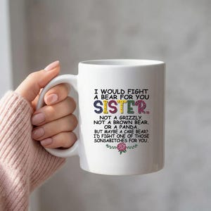 May include: White ceramic mug with a handle, featuring the text "I would fight a bear for you sister." The word "sister" is in colorful letters. The mug has a floral design and is held by a person.