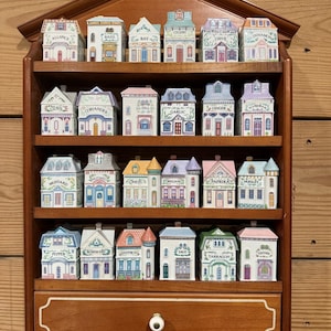 Op de afbeelding: Een houten kruidenrek met een lade, met 24 keramische kruidenpotjes in de vorm van kleurrijke huizen. Elk potje is gelabeld met een kruidennaam. Het rek is gemaakt van donker hout en gemonteerd op een lichtbruine houten muur.