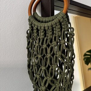 May include: An olive green macrame bag featuring a knotted net design and two circular wooden handles. The bag is constructed from thick cord, creating an open-weave pattern that extends from the wooden rings down to a series of small tassels.