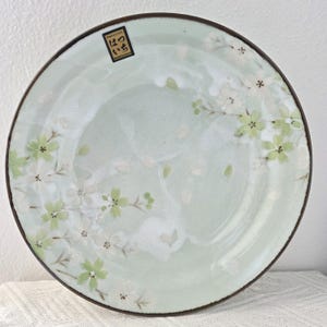 May include: A round, pale green ceramic plate with a brown rim. The plate is decorated with white and light green floral designs. The plate has a Japanese label in the upper left corner.