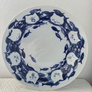 May include: White ceramic plate with a blue and white cat and fish design. The plate features a ring of stylized white cat faces and blue fish, creating a whimsical border around the central white space. A unique decorative piece.