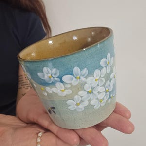 May include: A ceramic cup with a light blue exterior, decorated with white floral patterns and yellow centres. The interior is a warm, earthy brown. The cup is held in a person's hands.