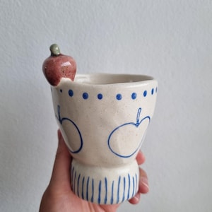 May include: A handcrafted ceramic cup with an apple motif. The cream-coloured cup has blue apple outlines, dots, and vertical lines. A small, red apple with a green stalk is attached to the rim. The cup is held by a person.