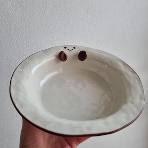 May include: A cream-coloured ceramic bowl with a brown rim and a smiling face design. The bowl features two brown circles for eyes and a curved line for a mouth. The bowl is held in a hand.