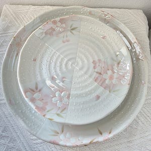 May include: Three white ceramic plates with a cherry blossom design, stacked on top of each other. The plates feature a light pink and green floral pattern. The plates are of varying sizes, with the largest at the base and the smallest at the top.