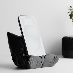 Könnte beinhalten: Ein schwarzer Telefonständer mit weichem, kissenartigem Design, der ein Smartphone hält. Der Ständer steht auf einer weißen Oberfläche, im Hintergrund eine kleine Topfpflanze. Das Telefon zeigt die Uhrzeit an.