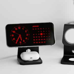 Könnte beinhalten: Schwarze Ladeständer für Telefon, Ohrhörer und Smartwatch. Das Telefon zeigt eine rote Uhr und einen Kalender für Januar an. Ein weißes Paar Ohrhörer liegt auf dem Telefonständer. Das Smartwatch-Ladegerät hat ein weißes Ladepad.