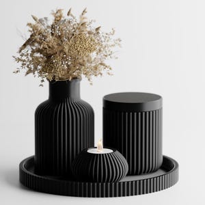 Könnte beinhalten: Ein schwarzes Dekorationsset mit einer Vase mit getrockneten Blumen, einem zylindrischen Behälter, einem kleinen Kerzenhalter mit einer brennenden Kerze und einem runden Tablett. Die Vase und der Behälter haben vertikale Rippendetails. Die Anordnung befindet sich vor einem weißen Hintergrund.