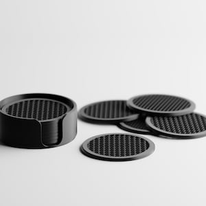 May include: A set of black round coasters with a textured surface, displayed with a holder. Several coasters are stacked in the holder, and a few are placed separately. The coasters have a woven pattern.