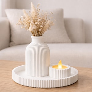 Puede incluir: Un jarr&oacute;n de cer&aacute;mica blanco y acanalado contiene flores secas, colocado sobre una bandeja a juego con una vela peque&ntilde;a. La vela tiene una llama amarilla. El arreglo est&aacute; sobre una mesa de madera frente a un sof&aacute; de tonos neutros.