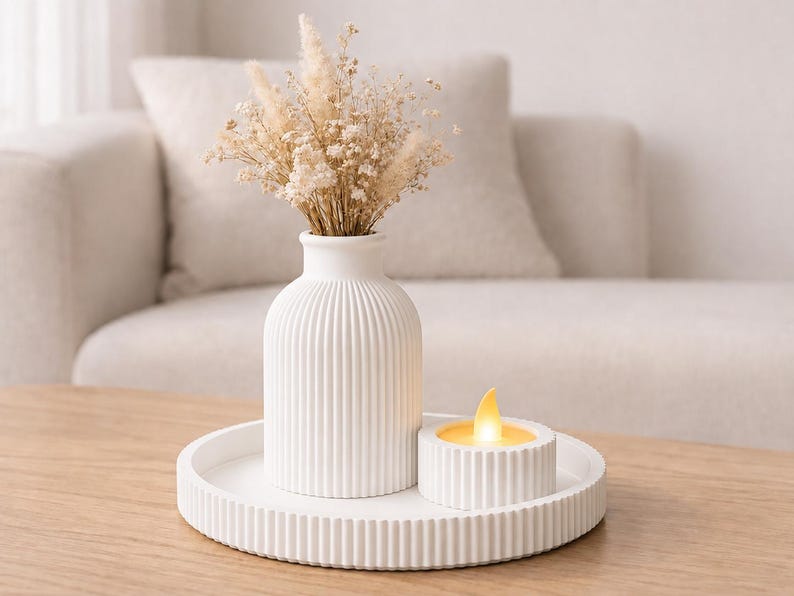 Puede incluir: Un jarr&oacute;n de cer&aacute;mica blanco y acanalado contiene flores secas, colocado sobre una bandeja a juego con una vela peque&ntilde;a. La vela tiene una llama amarilla. El arreglo est&aacute; sobre una mesa de madera frente a un sof&aacute; de tonos neutros.