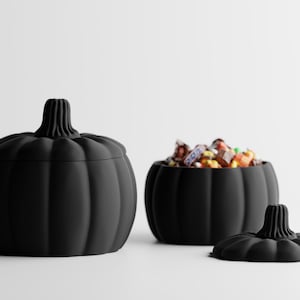 Peut inclure: Deux contenants noirs mats en forme de citrouille avec couvercles. Un contenant est fermé, tandis que l'autre est ouvert et rempli de bonbons emballés colorés, avec son couvercle assorti en forme de citrouille posé à côté sur la surface.