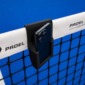 Könnte beinhalten: Eine schwarze Smartphone-Halterung aus Kunststoff, die an einem weißen Padel-Netzband befestigt ist. Das Band trägt den schwarzen Schriftzug PADEL und PADEL PRO COURT. Ein dunkelgraues Smartphone mit Dreifach-Kamerasystem steckt in der Halterung.
