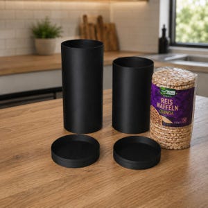 Op de afbeelding: Twee matzwarte cilindrische voorraadbussen met bijpassende deksels, de een hoger dan de andere, naast een doorzichtige plastic verpakking met ronde rijstwafels met het label REWE Bio Reis Waffeln Quinoa en een gewicht van 100g.