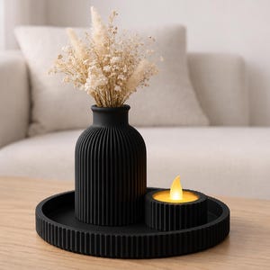 Puede incluir: Un conjunto decorativo negro sobre una mesa de madera. El conjunto incluye un jarr&oacute;n acanalado con flores secas, un peque&ntilde;o portavelas con una vela encendida y una bandeja redonda. El fondo presenta un sof&aacute; blanco.
