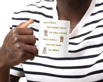 May include: White ceramic mug with the text "RICH PEOPLE HAVE BRAND LABLES ON THEIR CLOTHES HAPPY PEOPLE HAVE DOG HAIRS ON THEIR'S" and illustrations of three different dog breeds. The mug is held by a person wearing a black and white striped shirt.