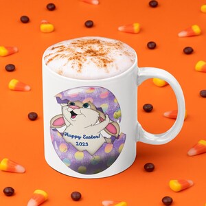 May include: White ceramic mug with a handle, featuring a cartoon bunny emerging from a cracked purple and blue egg. The mug has the text "Happy Easter! 2023". The mug is filled with a cappuccino, sprinkled with cinnamon, and set on an orange surface with candy corn.