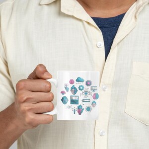 Könnte beinhalten: Weiße Keramik-Tasse mit einer Ansammlung farbenfroher Tech-Icons, darunter Gehirne, Wolken und Leiterplatten. Das Design ist in Blau-, Rosa- und Türkistönen gehalten. Die Tasse wird von einer Person gehalten, die ein hellgelbes Hemd mit Knöpfen trägt.