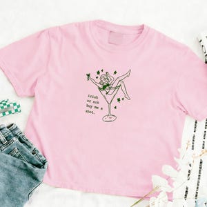 Könnte beinhalten: Hellrosa T-Shirt mit einer grünen Grafik einer Person in einem Martini-Glas, umgeben von Kleeblättern. Der Text "irish or not buy me a shot" steht darunter. Das Shirt ist aus weichem Material.
