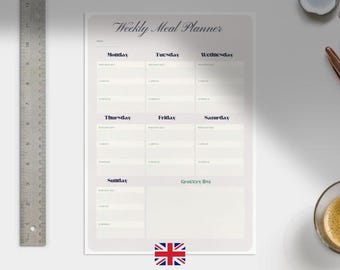 May include: A weekly meal planner with a light beige background and dark blue text. The planner includes sections for each day of the week and a grocery list. A metal ruler is on the left. A cup of coffee and a pen are on the right.