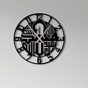 Op de afbeelding: Zwarte metalen wandklok met Arabisch kalligrafieontwerp. De klok heeft een cirkelvormig frame met Arabische cijfers en ingewikkelde kalligrafie in het midden. De wijzers zijn zilverkleurig. Aan een witte muur boven een witte bank.