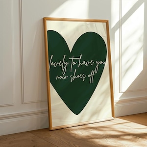 Peut inclure: Une affiche encadrée avec un grand cœur vert foncé sur un fond crème. Le cœur contient le texte blanc en écriture cursive : "lovely to have you now shoes off". Le cadre est de couleur marron clair.