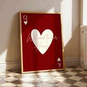 May include: A framed art print with a playing card theme. The red background features a white heart with the cursive text "Welcome to our House". The Queen and King of Hearts symbols are in the corners. The frame is a light wood colour.
