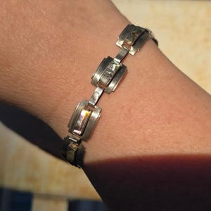 Könnte beinhalten: Ein silberfarbenes Metallgliederarmband mit rechteckigen Segmenten, die jeweils mit einem rechteckigen Stück irisierendem Abalone-Muschelmaterial eingelegt sind. Das Armband besteht aus geometrischen Metallrahmen, die durch flache Glieder verbunden sind.