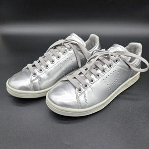 May include: A pair of metallic silver sneakers featuring a low-top design, matching grey laces, and a perforated letter R on the side panels. The shoes have a rounded toe, a padded collar, and a solid off-white rubber sole for a clean, uniform look.