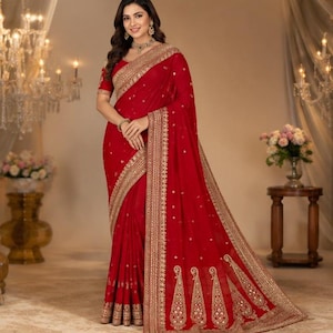 May include: A vibrant red saree featuring intricate gold embroidery along the borders and hem. The fabric is adorned with small, scattered gold embellishments. The garment includes a matching short-sleeved blouse with detailed gold trim at the sleeves.