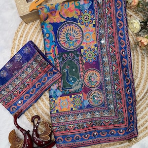 May include: A vibrant blue fabric piece featuring a multi-colored peacock and floral pattern with intricate geometric borders. The textile is embellished with small reflective mirror accents throughout the design, creating a detailed and ornate appearance.