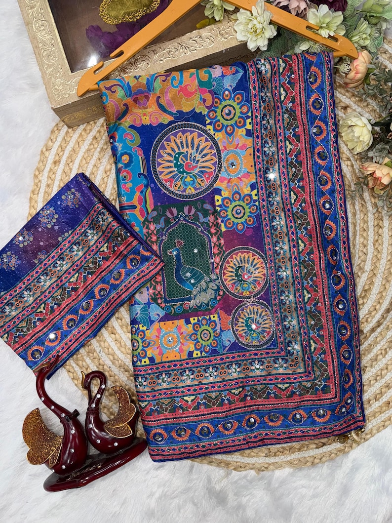 May include: A vibrant blue fabric piece featuring a multi-colored peacock and floral pattern with intricate geometric borders. The textile is embellished with small reflective mirror accents throughout the design, creating a detailed and ornate appearance.