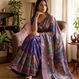 May include: A deep blue and purple saree featuring a vibrant, multi-coloured paisley and floral pattern throughout the fabric. The garment includes a wide, ornate border with intricate detailing and small reflective accents that catch the light.