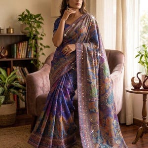 May include: A deep blue saree featuring a multi-colored paisley and floral pattern throughout the fabric. The garment is adorned with a wide border embellished with reflective circular mirror work and intricate embroidery along the edges.