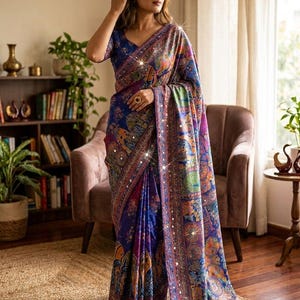May include: A deep blue saree featuring a vibrant, multi-colored printed pattern of traditional motifs and circular designs. The garment is accented with small, reflective mirror embellishments along the border and throughout the fabric.