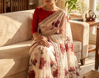 May include: A woman wearing a red long-sleeve blouse and a cream-colored sari with a floral pattern. The sari has a red and gold border. The woman is sitting on a beige sofa in a well-lit room.